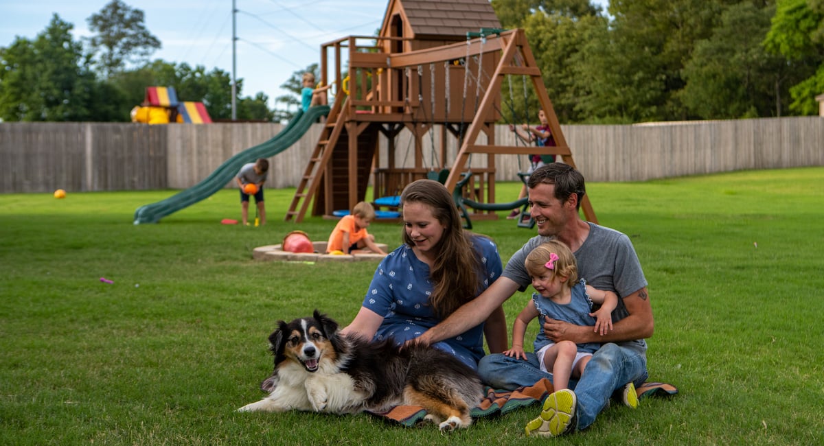 family sits on grass and plays in backyard with dog 